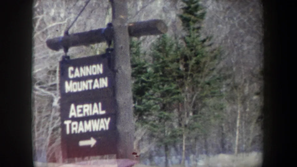 1962 8mm close view of the black wooden Cannon Mountain Aerial Tramway sign in a snowy forest setting.