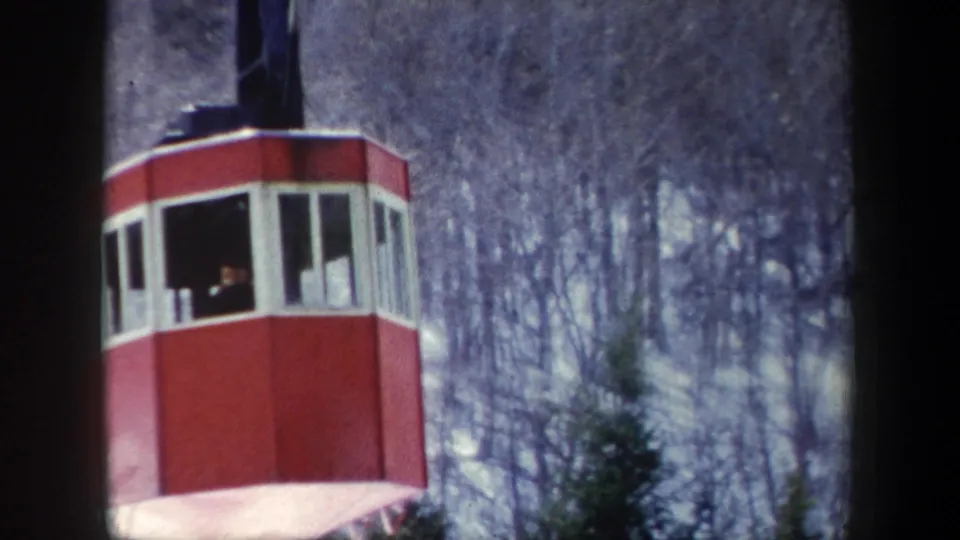 1962 8mm frame showing a red-and-white aerial tram car suspended above winter trees with small passengers inside.