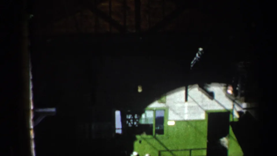 8mm frame showing a green cableway station building beneath a dark structure, with railings and mechanical elements visible.