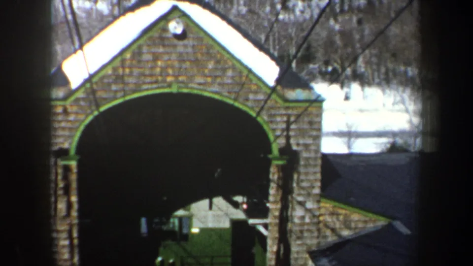 1962 New York frame of a gabled arched stone structure, its roof edged with snow and bare trees visible beyond.