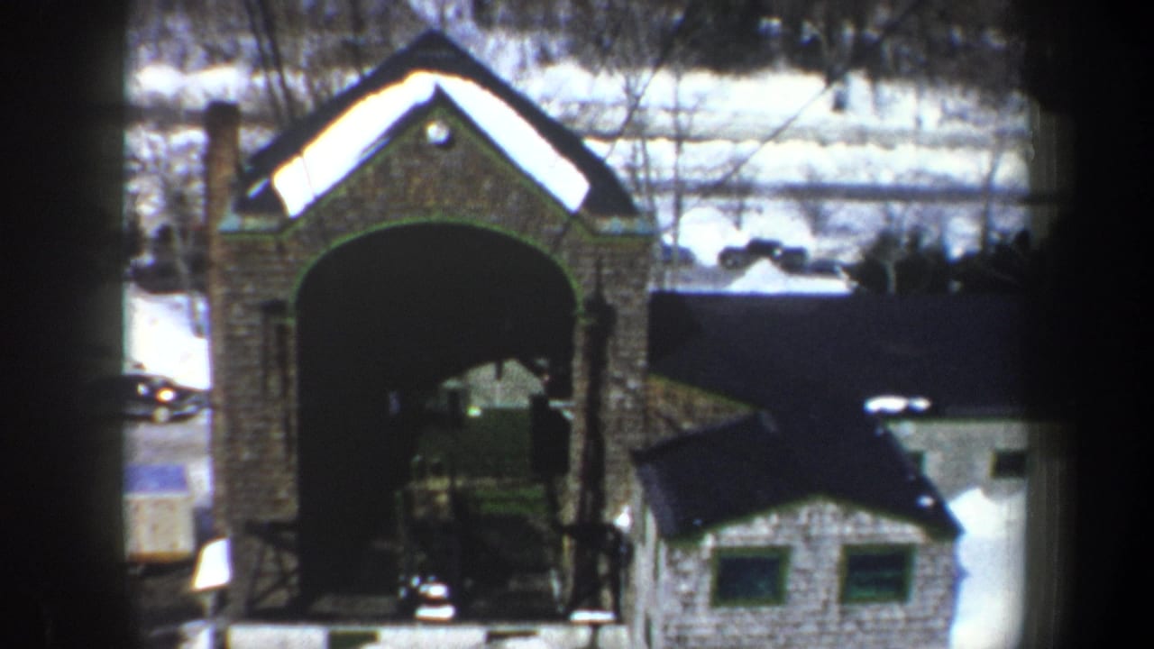 1962 New York 8mm frame showing a snow-covered stone archway, nearby small building, road, and distant parked cars.