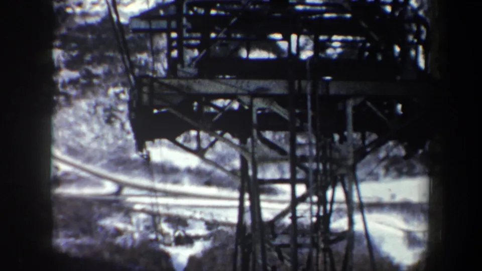 Aerial-feeling 1962 New York view of a utility tower structure, cables, snow-covered road, and wooded winter hillside.