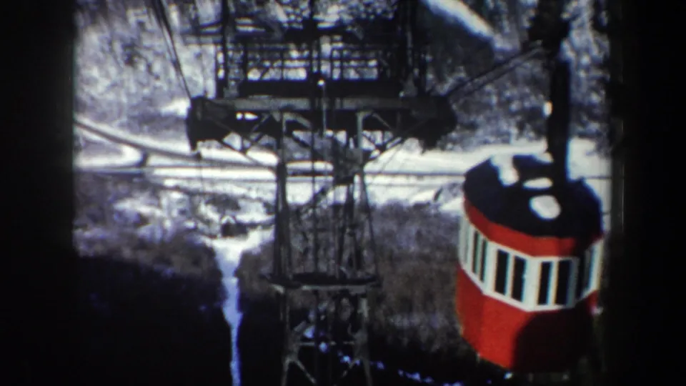 Red-and-white cable car descends past a tall metal support tower in a snowy New York mountain landscape.