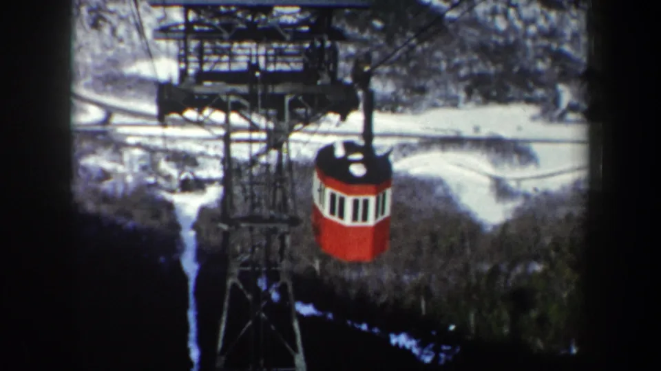 1962 home-movie frame of a red cable car centered beneath a steel tower against snow-dusted New York woods.