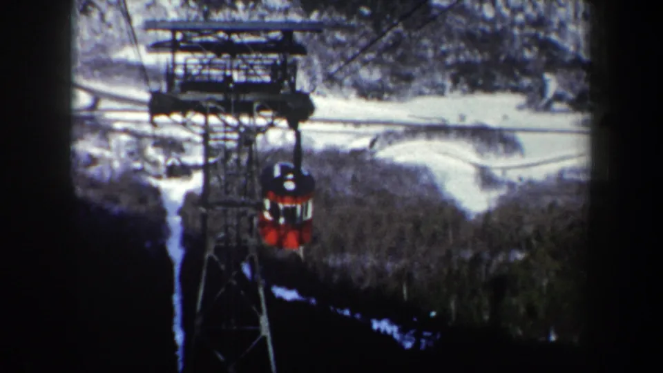 Red cable car appears smaller as it descends along the cable line beyond the steel tower in New York mountain terrain.