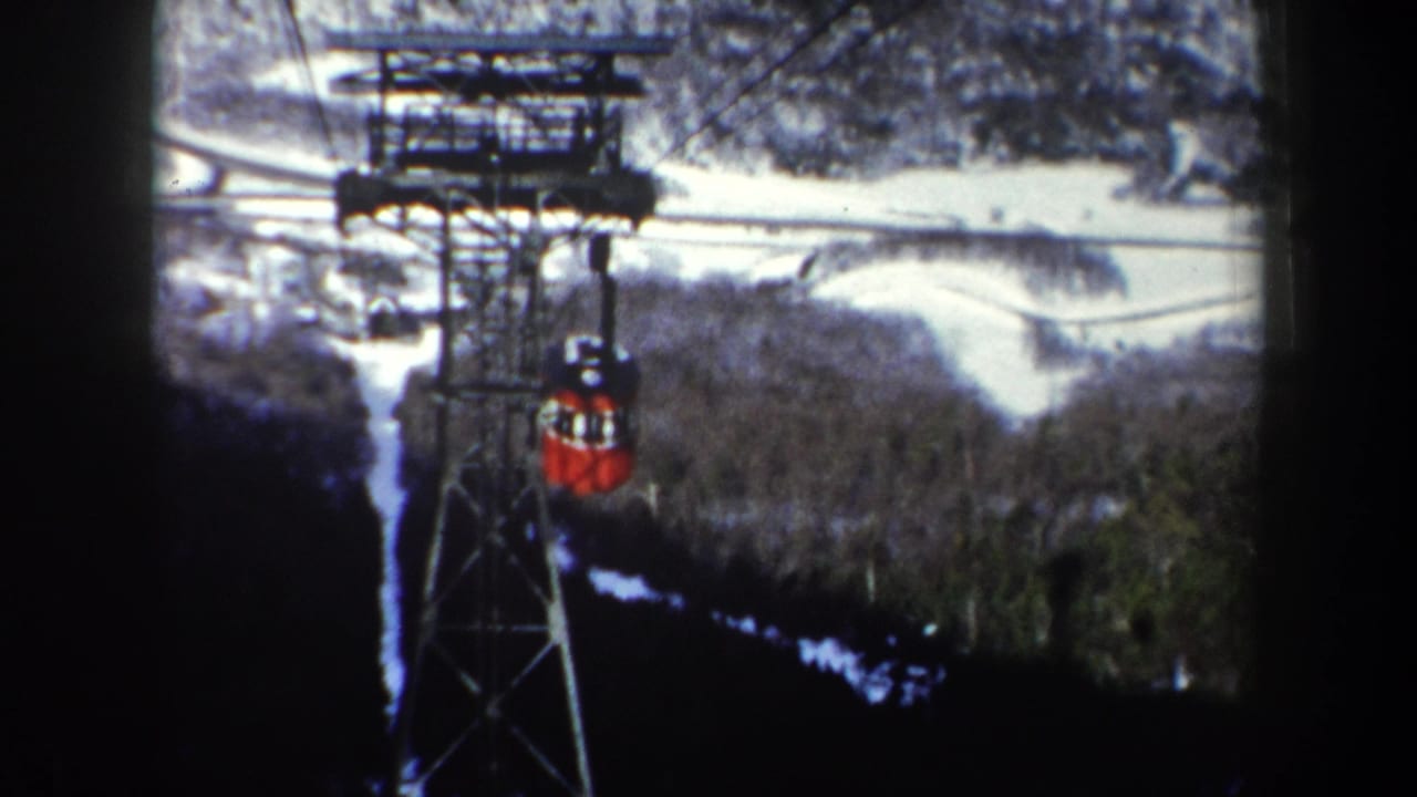 1962 New York 8mm frame showing a red tram cabin near a support tower above snow-covered forest and winding pale terrain.