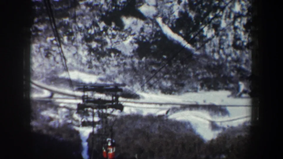 1962 New York 8mm frame with a red tram cabin below overhead cables, seen against snow-covered mountain ground and trees.