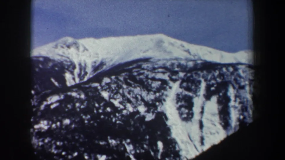 1962 New York winter landscape frame showing a snow-covered mountain ridge beneath clear blue sky and darker forested slopes.