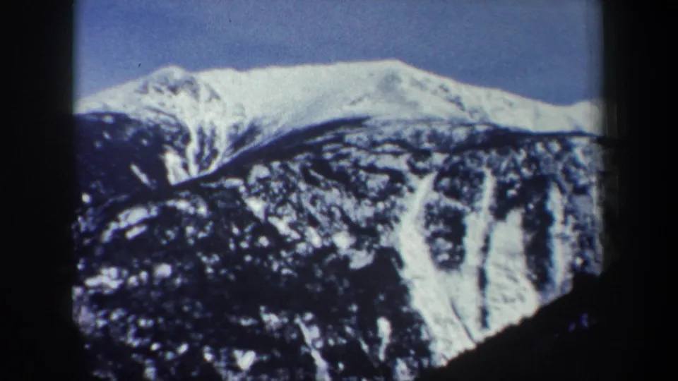 1962 New York 8mm frame with a broad snowy ridge above a dark wooded mountain slope and blue sky.