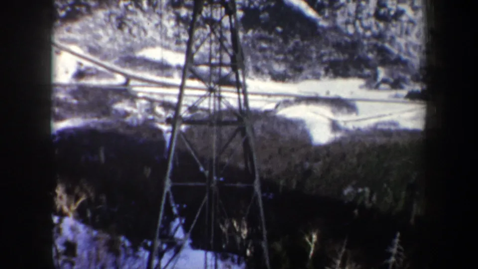 A 1962 New York mountain lift tower stands beneath cables, with snow patches, hillside terrain, and dark cabin-edge framing visible.