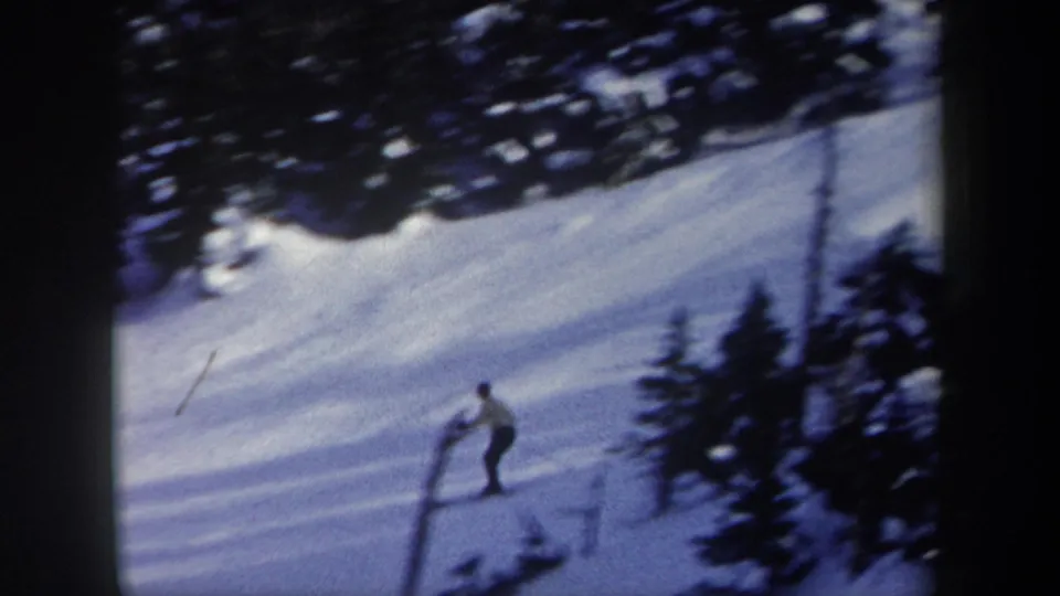 1962 New York 8mm frame of a distant skier in a light jacket moving down a groomed snowy run beside trees.