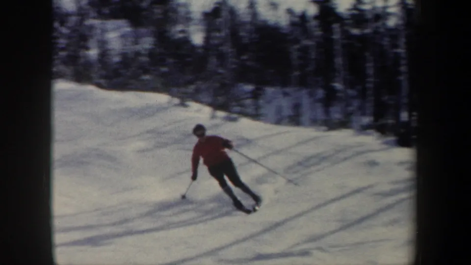 A skier in a red jacket and dark pants moves across a snowy New York slope, with trees and long shadows in the background.