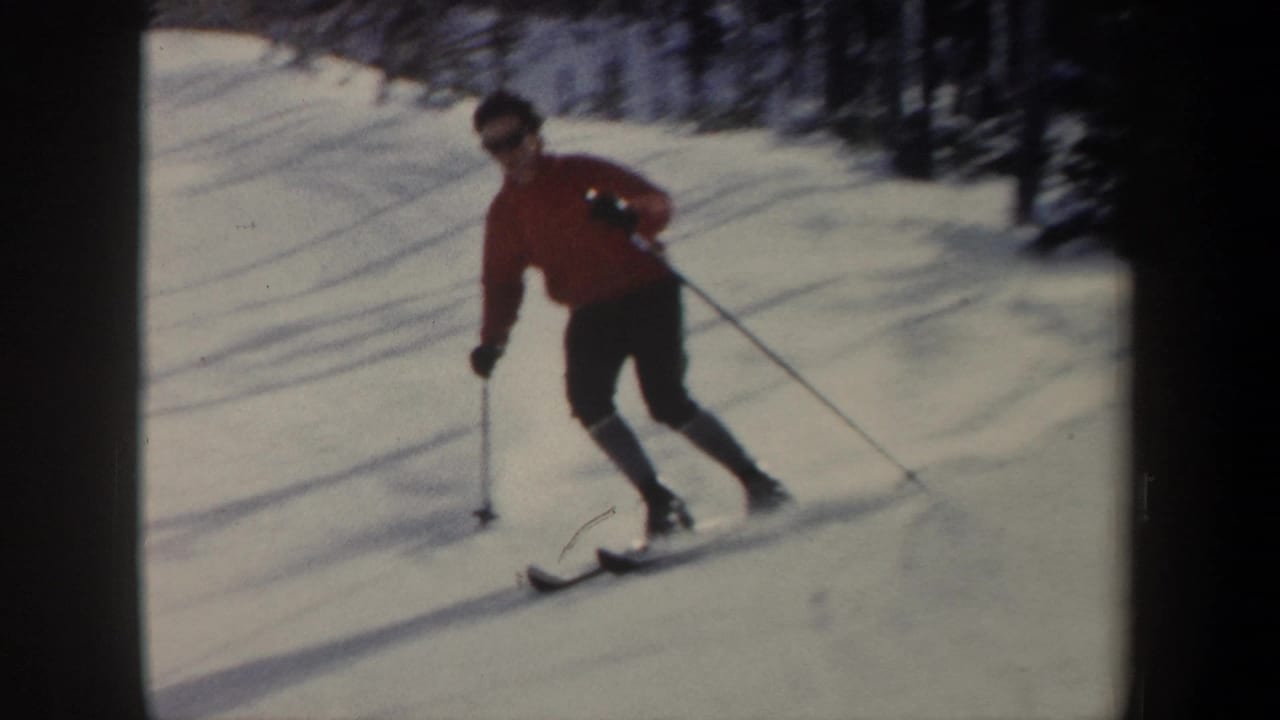 Skier in a red jacket turns across a snowy slope with trees behind.