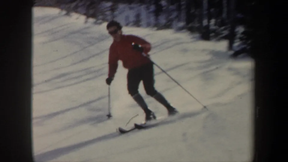 1962 New York home-movie frame showing a skier in a red jacket and dark pants using poles on a snowy wooded slope.