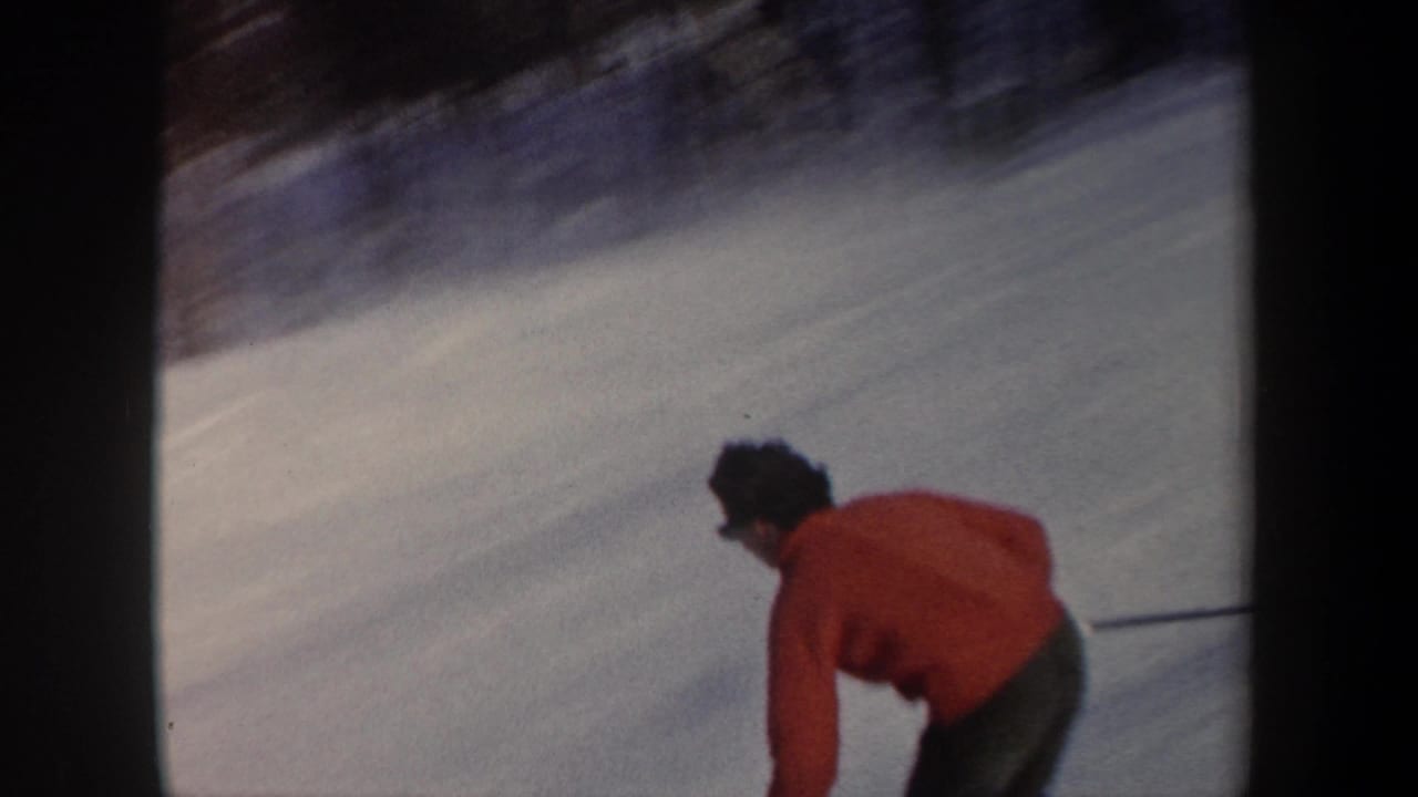 Home-movie frame from the New York ski clip showing the red-jacketed skier low in the frame on a snowy downhill slope.