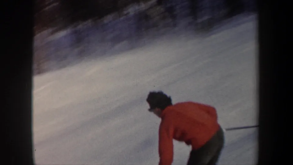 Home-movie frame from the New York ski clip showing the red-jacketed skier low in the frame on a snowy downhill slope.