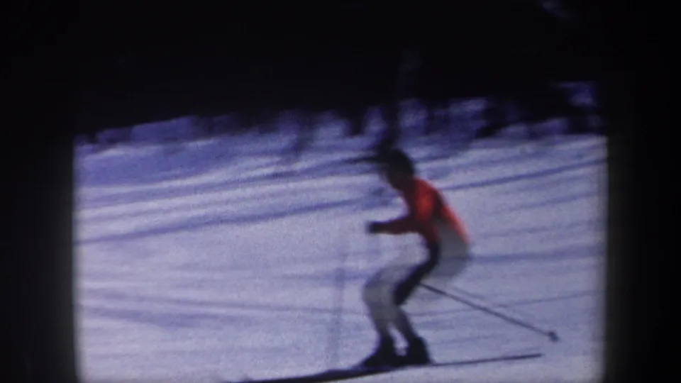 A skier in an orange jacket and dark pants crosses a snowy slope with ski poles in 1962 New York home-movie footage.