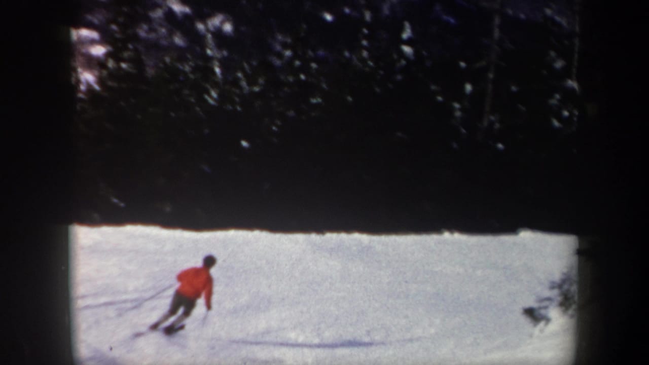 1962 New York 8mm frame showing a red-jacketed skier carving across a snowy slope near evergreens.