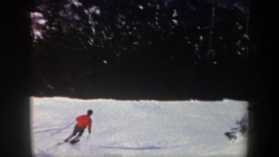 1962 New York 8mm frame showing a red-jacketed skier carving across a snowy slope near evergreens.