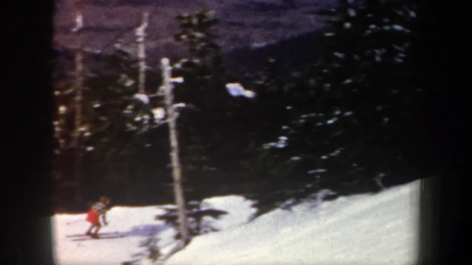 1962 New York 8mm frame showing a red-jacketed skier descending near evergreens and a pole on a snowy run.