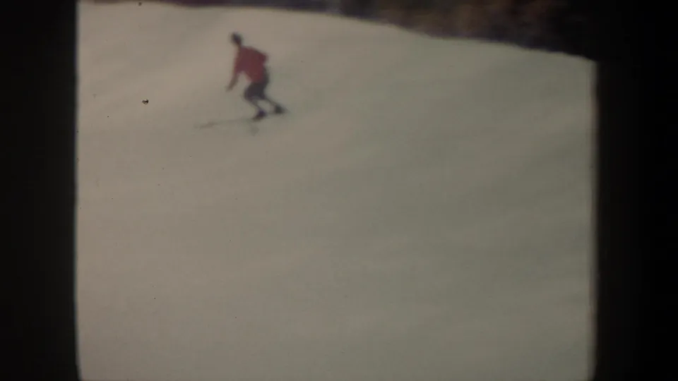 The skier in a red jacket leans forward while turning on the snowy slope, seen in soft 1962 8mm footage from New York.