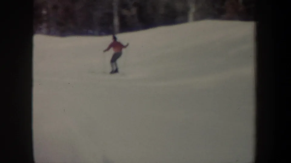 A skier in a red jacket continues downhill across a pale snowfield, with dark trees beyond and visible 8mm frame edges.