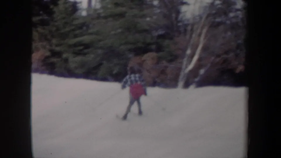 1962 New York 8mm frame of a skier in plaid and a red pack moving across a wooded snowfield.