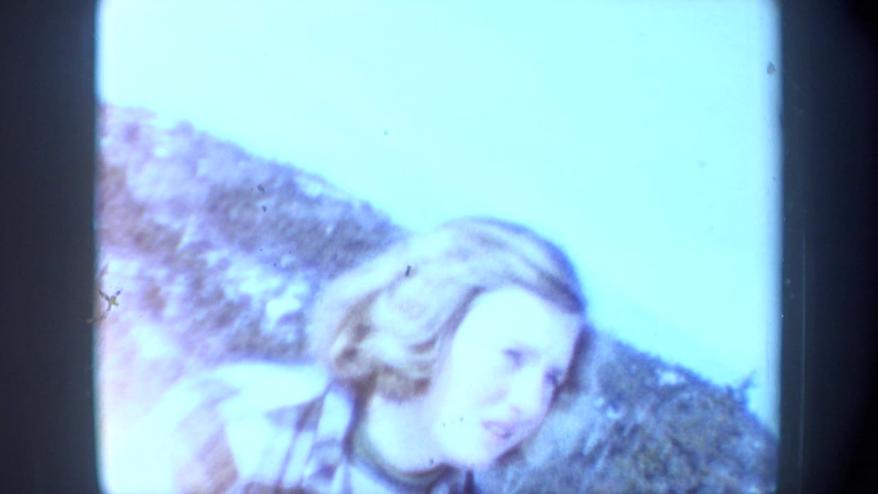 1962 New York 8mm frame of a blonde woman in plaid leaning near a snowy hillside, softened by bright exposure and film fade.