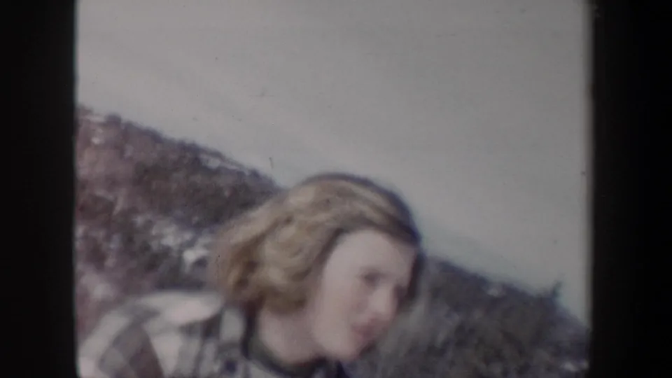 Close home-movie view of the blonde woman in plaid outdoors, with snow-dusted terrain and muted 8mm color.