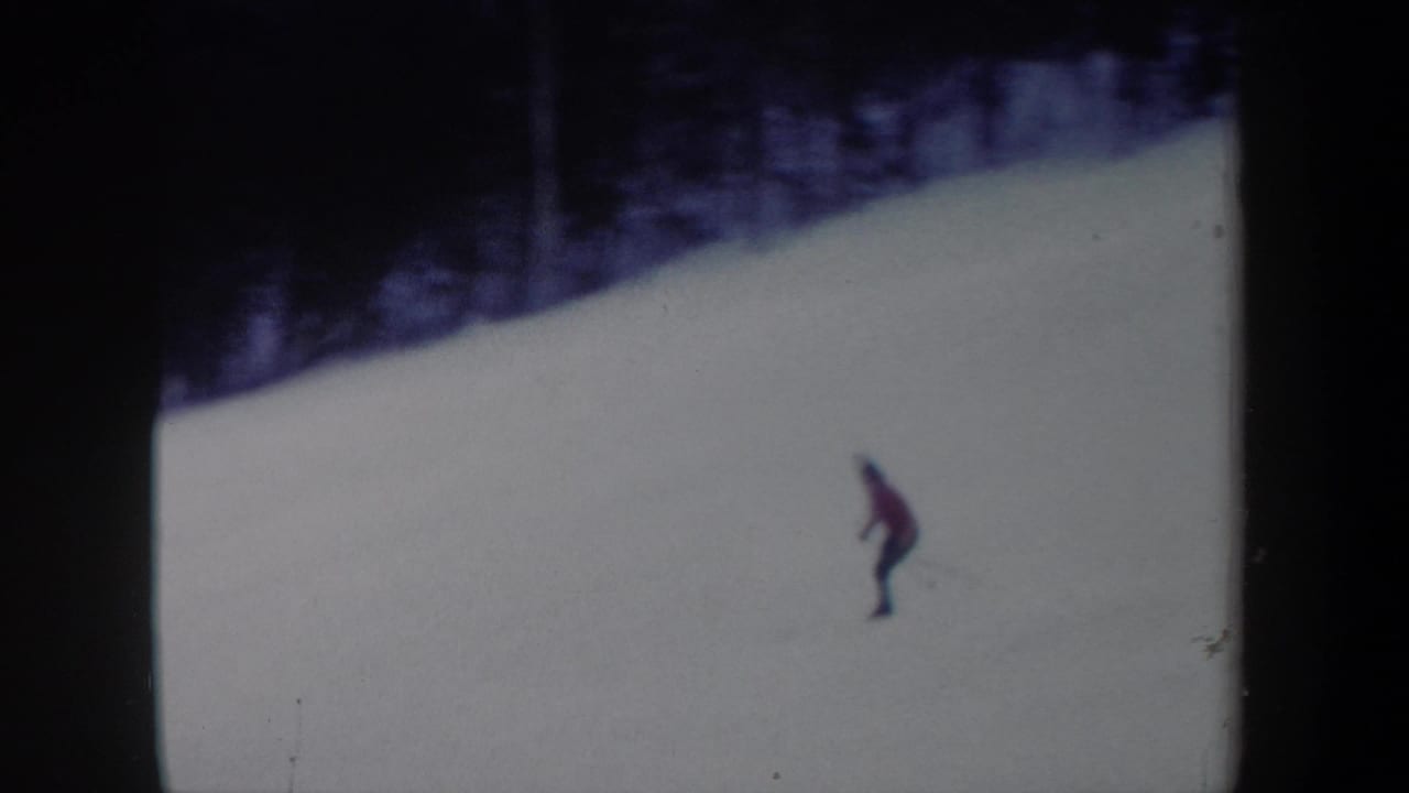 1962 New York 8mm frame showing a distant skier in red moving down a snowy slope with dark evergreen trees above.