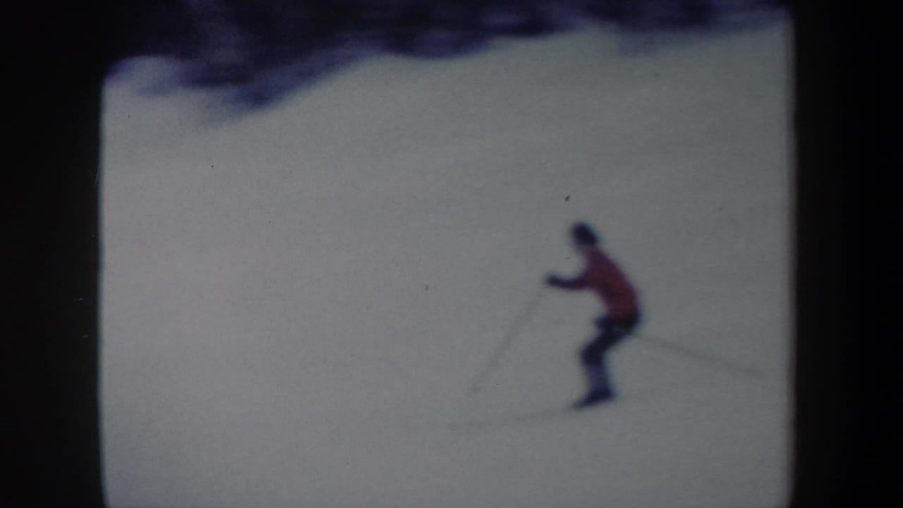 A lone skier in red and dark pants crosses the snowy slope in soft 8mm color, with ski poles visible.