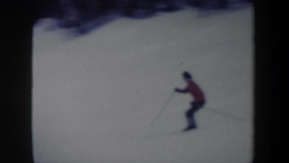 A lone skier in red and dark pants crosses the snowy slope in soft 8mm color, with ski poles visible.
