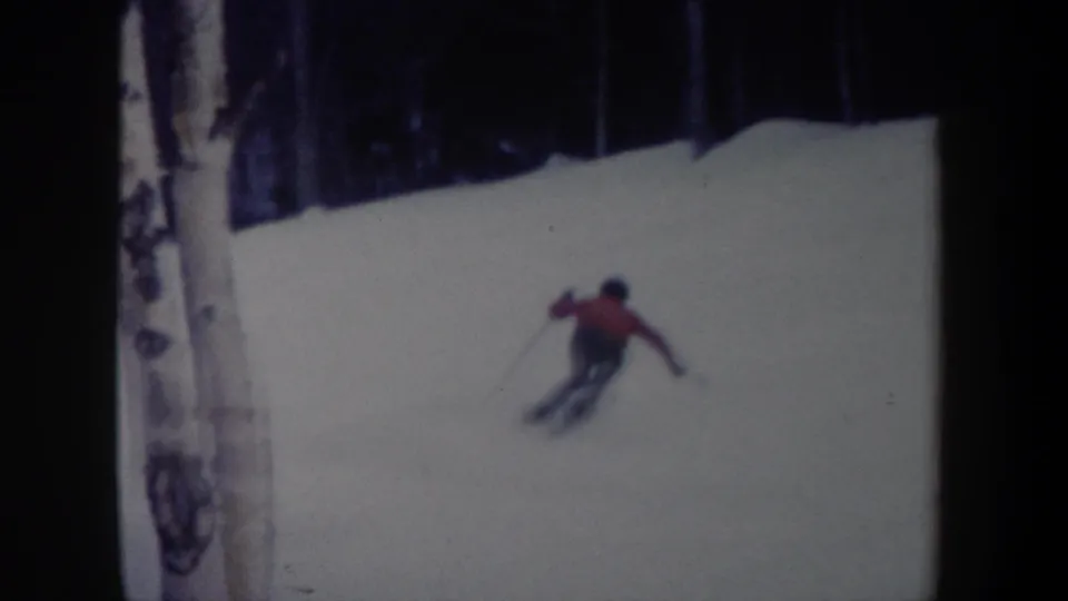 The skier in a red jacket leans through a controlled turn on the snow, with trees and dark film edges nearby.