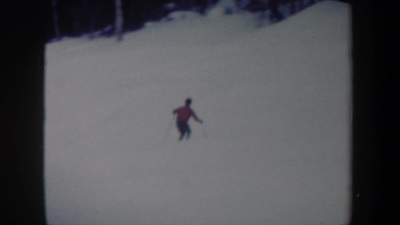 1962 New York 8mm frame showing a lone skier in red moving downhill across a pale snowy slope, with bare trees along the top edge.