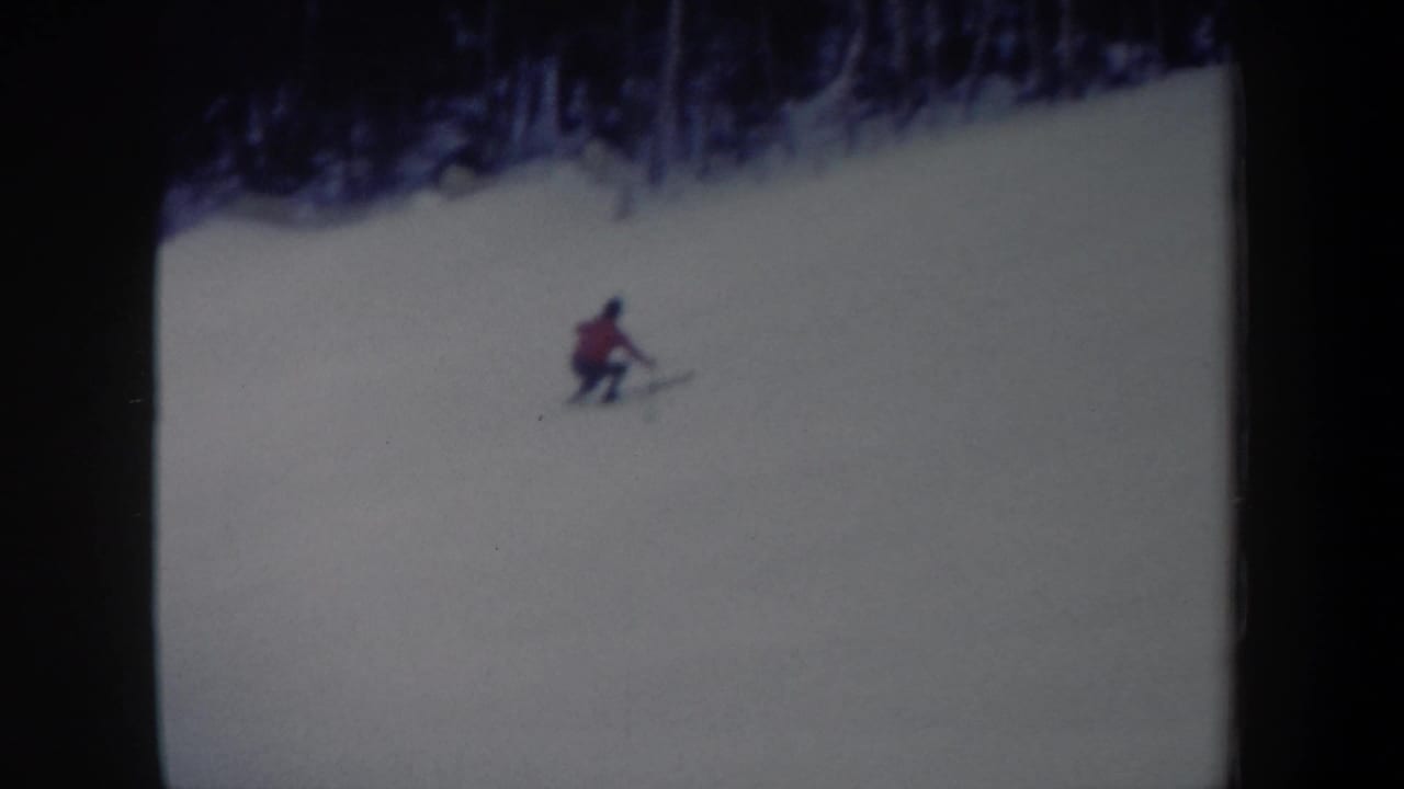 1962 New York home-movie frame with a distant skier in a red jacket turning on a wide snowy slope beneath dark winter trees.