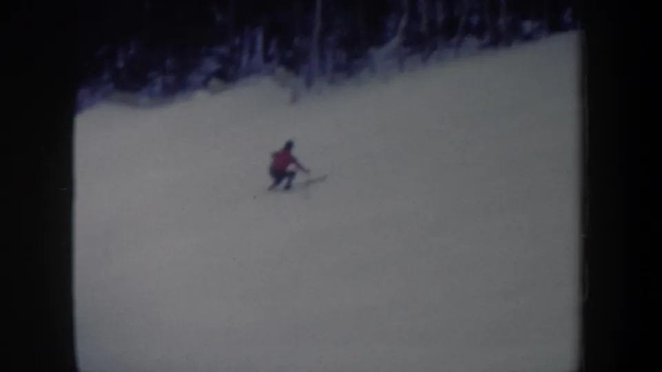 1962 New York home-movie frame with a distant skier in a red jacket turning on a wide snowy slope beneath dark winter trees.