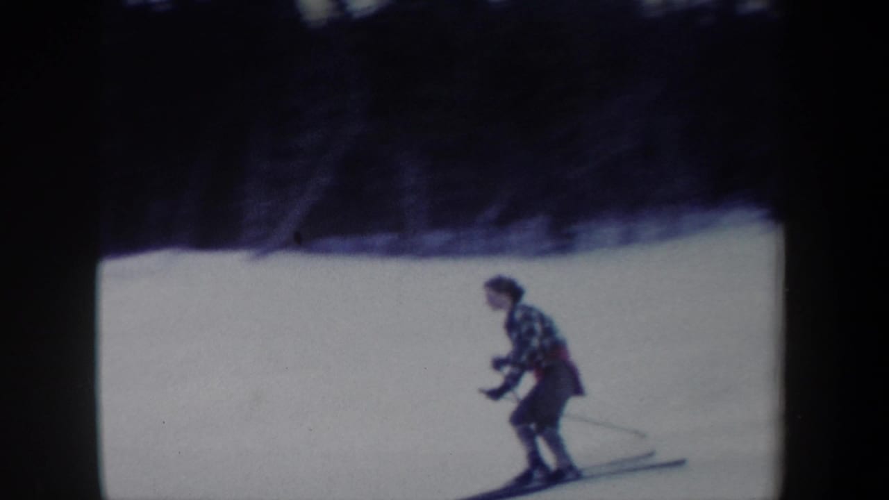 1962 New York 8mm frame showing a woman in a plaid jacket skiing downhill with poles on a snow-covered slope.