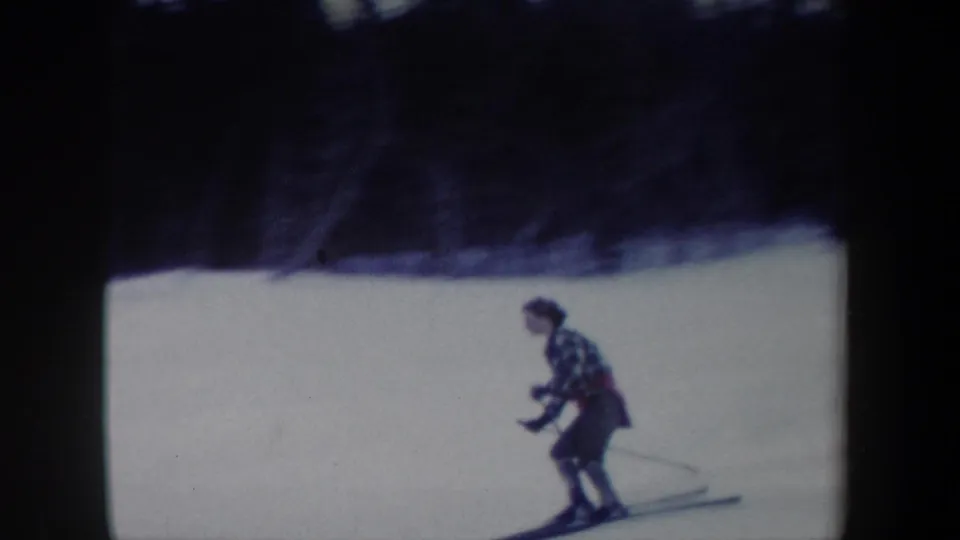 1962 New York 8mm frame showing a woman in a plaid jacket skiing downhill with poles on a snow-covered slope.