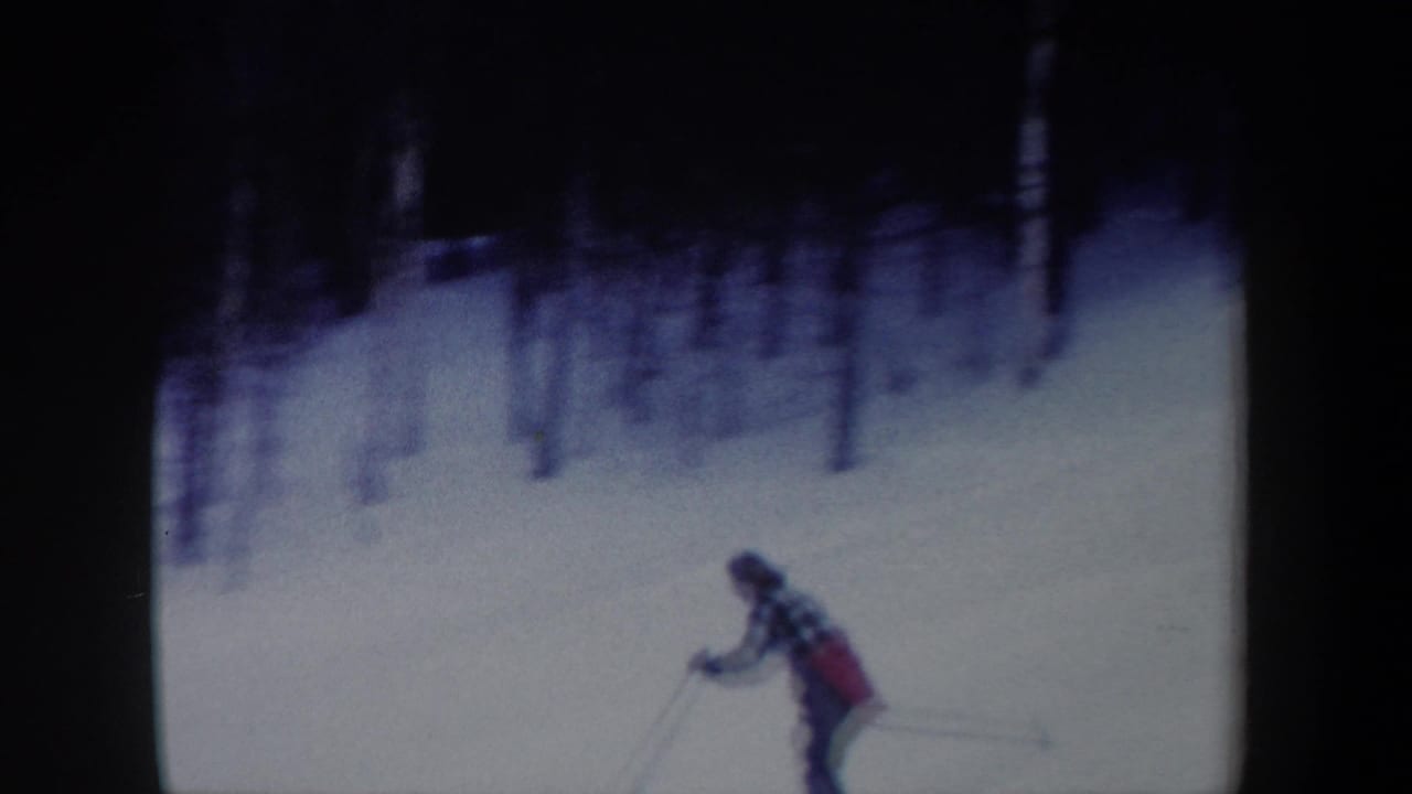1962 New York home-movie frame of a woman skier moving across a snowy mountain slope beneath a dark wooded edge.