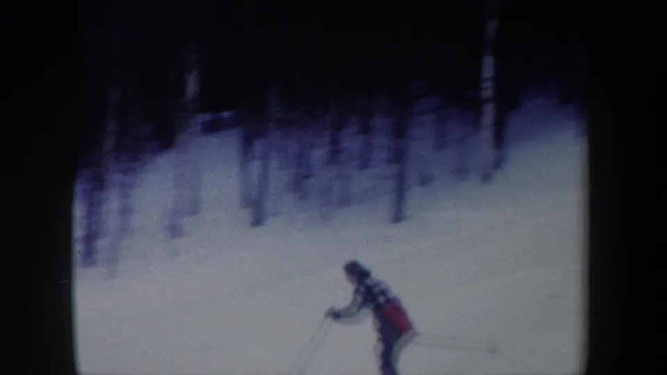 1962 New York home-movie frame of a woman skier moving across a snowy mountain slope beneath a dark wooded edge.