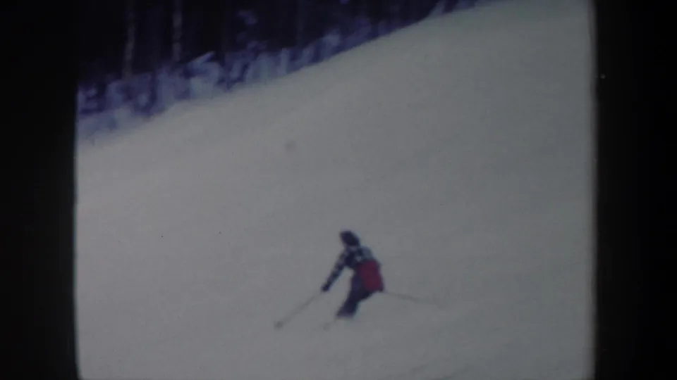 1962 New York home-movie frame of a woman skier turning on a broad snowy slope, with a dark forest edge above.