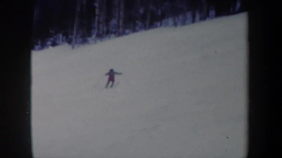 Distant skier in 1962 New York 8mm footage traversing a snow-covered hillside below bare trees.