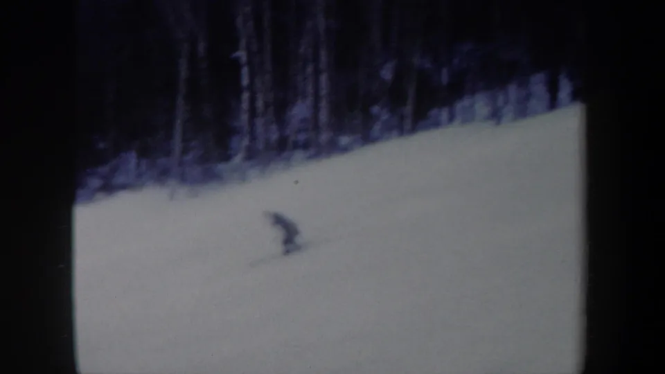 The skier appears lower in the frame and bent forward while crossing the snowy hillside in vintage 8mm footage.
