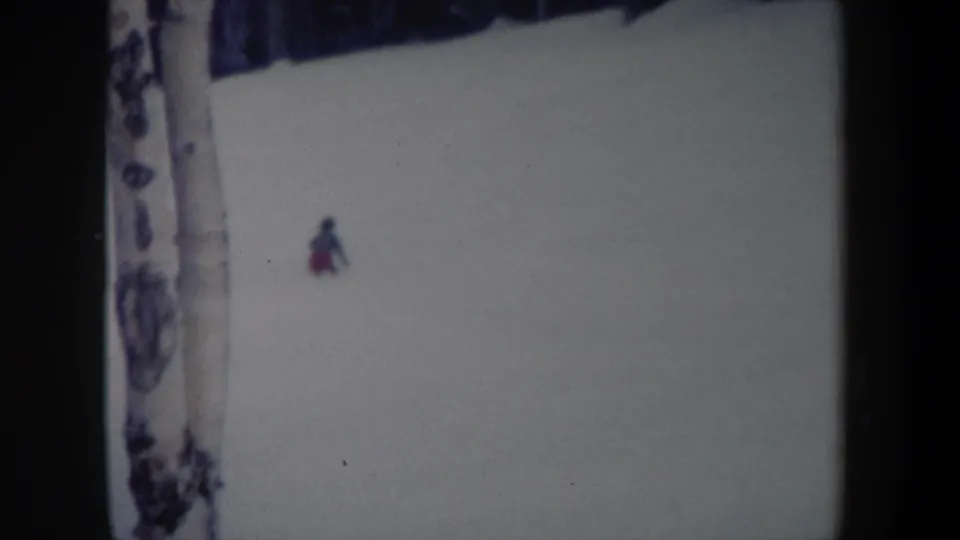 1962 New York home-movie frame showing a lone skier descending near a foreground birch trunk.