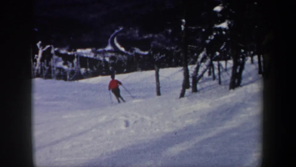 1962 New York 8mm frame with the skier turning through snow near a line of dark bare trees.