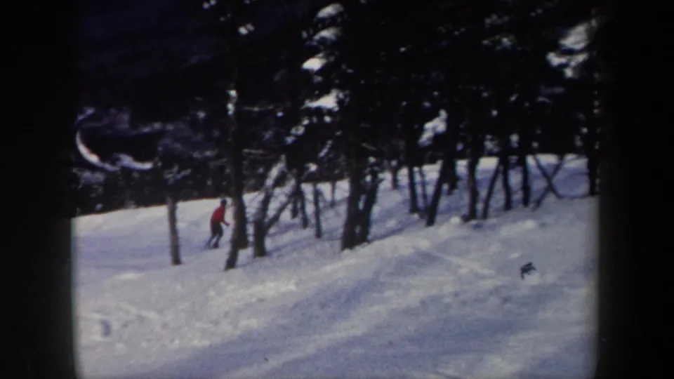 1962 New York 8mm frame showing the skier lower on the wooded snowy slope, partially framed by tree trunks.