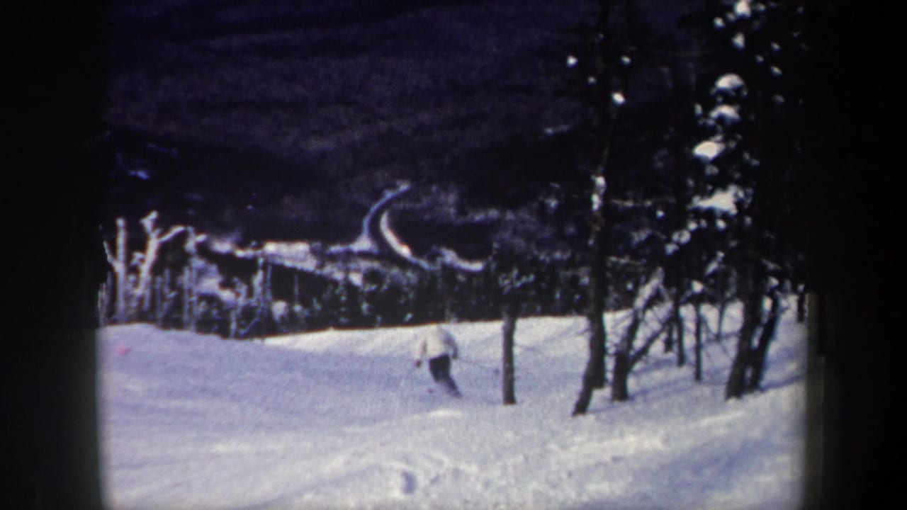 1962 New York 8mm frame showing a distant skier descending a snowy mountain slope with evergreen trees and a curved run in view.