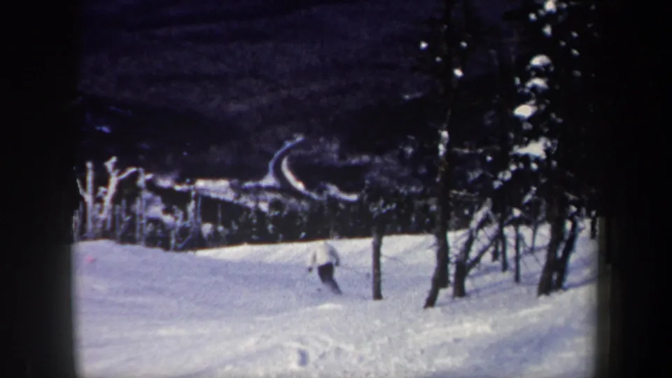 1962 New York 8mm frame showing a distant skier descending a snowy mountain slope with evergreen trees and a curved run in view.