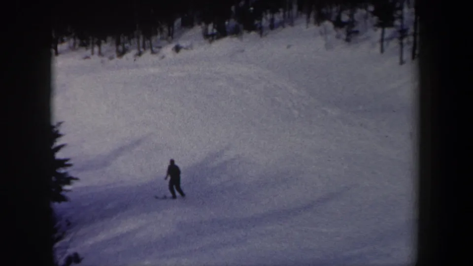 1962 New York 8mm frame of a lone skier moving down a snowy run, framed by evergreen trees and shaded snow.