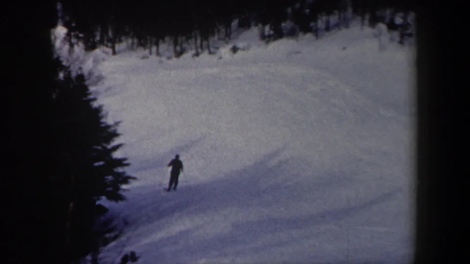 In 1962 New York 8mm footage, a lone skier appears small on a broad snowy slope with evergreen trees and heavy shadow along the run.